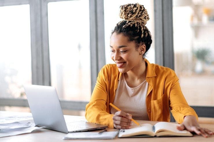 Young woman smiling as she takes notes beside her laptop